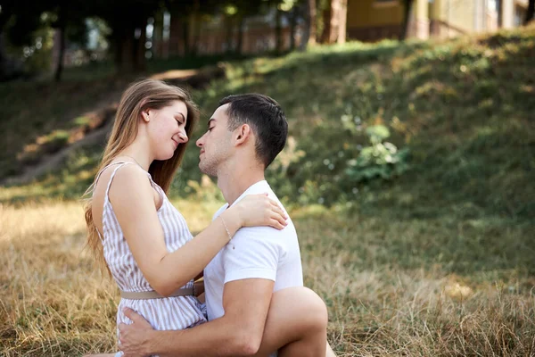 Young couple in love sitting on green yellow grass lawn hugging embracing kissing. Blond woman wearing stripy short overall and brunette man in white t-shirt blue shorts on romantic date. Relationship