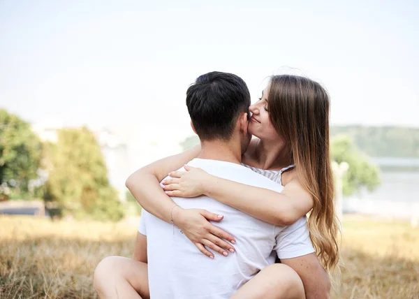 年轻夫妇坐在翠绿的黄草草坪上拥抱亲吻 在浪漫的约会中 金发女人总体上穿着条纹短裙 黑发男人穿着白色T恤蓝色短裤 — 图库照片