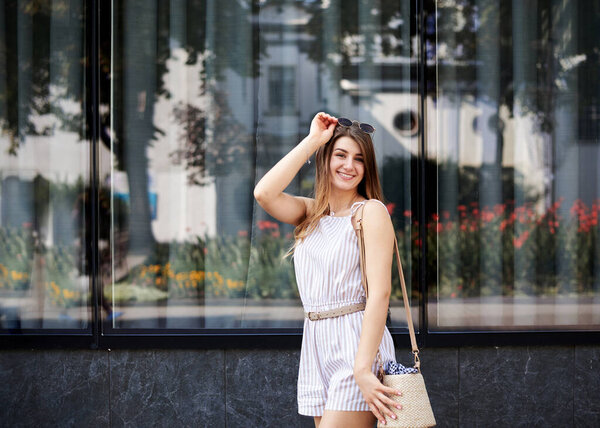 Молодая блондинка в белых полосатых коротких очках и солнечных очках, стоящая рядом с большим стеклянным зданием. Три четверти портрета худенькой девушки, позирующей для фото на курорте. Отдых в отпуске
