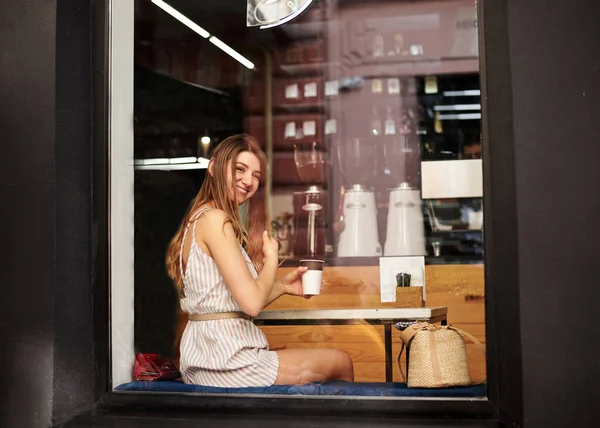 Young blond woman, wearing white overall, sitting by huge glass window in coffee shop. Pretty girl on dinner break, drinking coffee tea cappuccino in street city cafe in summer.