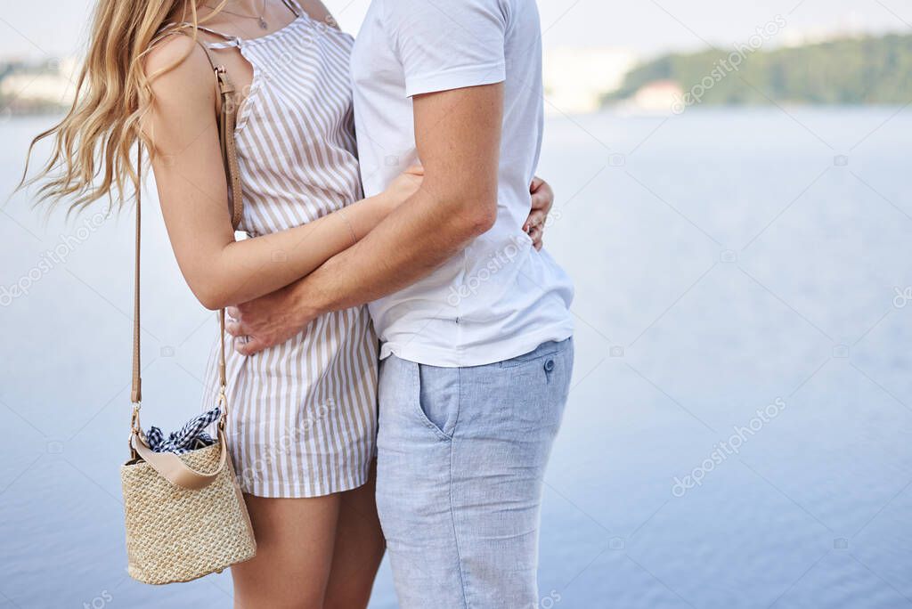 Man, wearing white t-shirt, blue shorts, is hugging woman in stripy summer overall. Bodies of young couple in love, embracing. Close-up picture of male and female body together in front of blue lake.