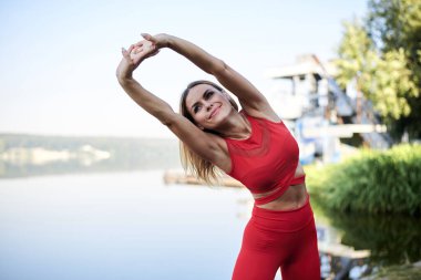 Genç sarışın kadın, kırmızı tayt giyiyor, spor ayakkabıları üstte ve beyaz, yazın göl kenarında sabah egzersizleri yapıyor. Fitness yoga esneme antrenmanı suyun yanında. Sağlıklı yaşam konsepti. Aktif yaşam biçimi
