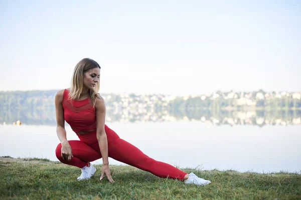 年轻的金发女人 穿着红色的腿 头皮和白色的运动鞋 夏天在湖边晨练 健身瑜伽在水的作用下伸展训练 健康生活的概念 积极的生活方式 — 图库照片