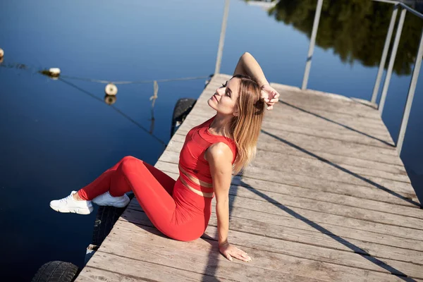 若いブロンドの女性は 夏の朝に木製の桟橋ドックに座って赤いフィットネス服を着ていた 健康的なアクティブライフコンセプト 街の外で練習した後に休んでいる女の子の肖像画 — ストック写真