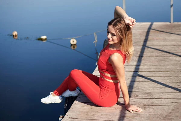 若いブロンドの女性は 夏の朝に木製の桟橋ドックに座って赤いフィットネス服を着ていた 健康的なアクティブライフコンセプト 街の外で練習した後に休んでいる女の子の肖像画 — ストック写真