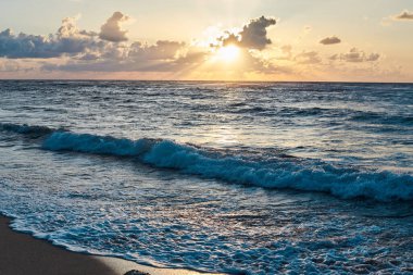 Denizde gün batımı. Akşamları okyanusta turkuaz su. Küçük bulutlu sarı gökyüzü ve deniz kenarındaki kumlu sahilde mavi dalgalar. Cennet tatil beldesi manzarası. Tropik tatil beldesi fotoğrafı..