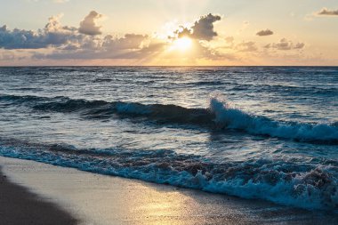 Denizde gün batımı. Akşamları okyanusta turkuaz su. Küçük bulutlu sarı gökyüzü ve deniz kenarındaki kumlu sahilde mavi dalgalar. Cennet tatil beldesi manzarası. Tropik tatil beldesi fotoğrafı..