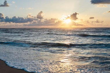 Denizde gün batımı. Akşamları okyanusta turkuaz su. Küçük bulutlu sarı gökyüzü ve deniz kenarındaki kumlu sahilde mavi dalgalar. Cennet tatil beldesi manzarası. Tropik tatil beldesi fotoğrafı..