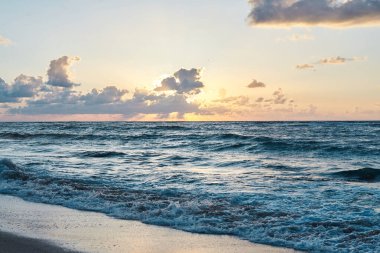 Kayalık denizde gün batımı. Akşamları okyanusta turkuaz su. Deniz kenarında küçük bulutlu sarı gökyüzü ve kumlu sahilde mavi dalgalar. Cennet tatil köyü manzarası. Okyanus dalgalarının yakın plan resmi.