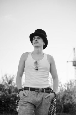 Beyaz bluzlu, siyah şapkalı, güneş gözlüklü ve düşünen bir gencin dörtte üçü. Terk edilmiş inşaat alanındaki yaratıcı adamın siyah beyaz fotoğrafı. Sanat Evi.