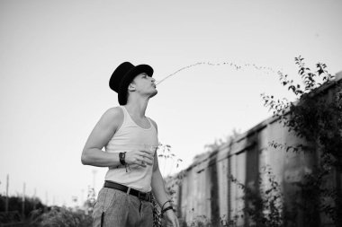 Gri pantolonlu, beyaz bluzlu, siyah şapkalı, su bardağı tutan ve yan tarafa tüküren bir gencin dörtte üçü portresi. Terk edilmiş şantiyedeki yaratıcı adamın siyah beyaz resmi.