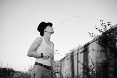 Gri pantolonlu, beyaz bluzlu, siyah şapkalı, su bardağı tutan ve yan tarafa tüküren bir gencin dörtte üçü portresi. Terk edilmiş şantiyedeki yaratıcı adamın siyah beyaz resmi.