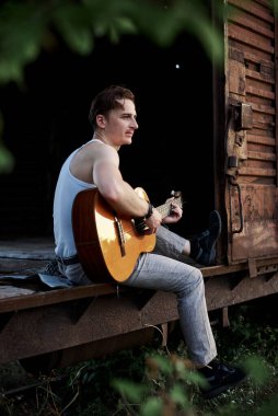 Beyaz tişört ve gri pantolon giyen yakışıklı genç adam, eski tren vagonunda oturuyor, gitar çalıyor, yazları terk edilmiş trende gitaristli esmer erkek müzisyen besteler yapıyor..