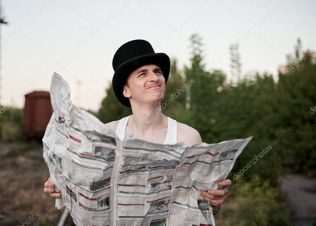Young man, wearing white t-shirt and black magician hat is holding newspaper, thinking, reading, smiling. Three-quarter portrait of magician with paper on abandoned industrial zone.