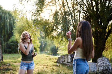 İki kız kardeş, parkta birbirlerine sabun köpüğü üflüyorlar, gülüyorlar, gülümsüyorlar, genç güzel kızlar, kot şort giyiyorlar, yeşil bej bluzlar giyiyorlar, dışarıda eğleniyorlar. İki kız arkadaşın çeyrek portresi..