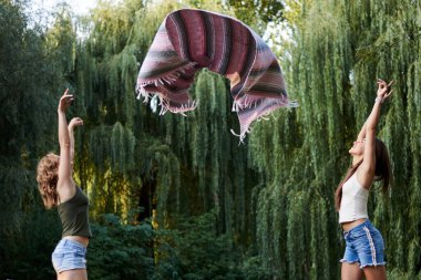 Yazın parkta havaya pembe ve gri Meksika battaniyesi atan iki kız kardeş. Genç kızlar, kot şort, bej ve haki rengi bluz giyiyorlar, eğleniyorlar. İki kız arkadaşın tam boy portresi..