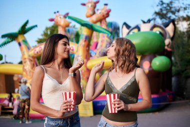 İki kız kardeş, eğlence parkında ayakta durmak, patlamış mısırı kağıt kutuda tutmak, gülümsemek, eğlenmek. Güzel kızlar. Kot pantolon, bej ve yeşil bluz giyiyor, lunaparkta dinleniyor.