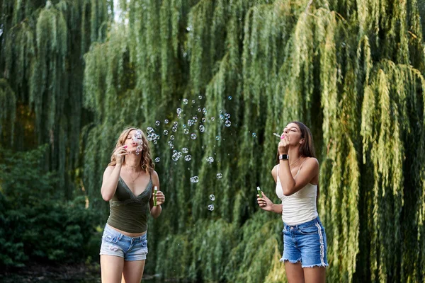 两个姐姐夏天在公园里吹肥皂泡年轻漂亮的女孩 穿着牛仔裤短裤和绿色米黄色上衣 在外面玩的很开心 正在度假的瘦女人的四分之三画像 — 图库照片