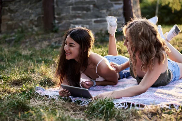 夏に公園の芝生の上に毛布の上に横たわる2人の姉妹は タブレットで楽しいビデオを見ています きれいな女の子 身に着けているライトブルーのジーンズのショートパンツと緑のベージュのトップ リラックスした外 お互いをくすぐる — ストック写真