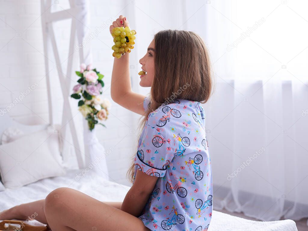 Young brunette women girl, wearing purple violet cotton pajamas, sitting on white bed in light room apartment, smiling, eating green grapes for breakfast.Wedding morning celebration. Sleepwear design