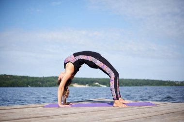 Çıplak ayaklı genç esmer kadın, siyah ve mor spor kıyafeti giyiyor, yaz aylarında ahşap iskelede mor yoga minderinde jimnastik köprü pozu veriyor. Formda kız, geriniyor ve göl kenarında yoga yapıyor.