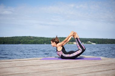 Çıplak ayaklı genç esmer kadın, siyah ve mor spor kıyafeti giyiyor, yaz aylarında tahta iskelede menekşe yoga minderinin üzerinde geriniyor. Göl kenarında yoga pozları veren formda bir kız. Sağlıklı yaşam tarzı.