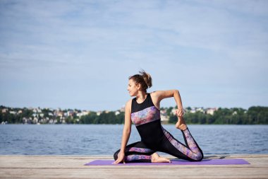 Çıplak ayaklı genç esmer kadın, siyah ve mor spor kıyafeti giyiyor, yaz aylarında tahta iskelede menekşe yoga minderinin üzerinde geriniyor. Göl kenarında yoga pozları veren formda bir kız. Sağlıklı yaşam tarzı.