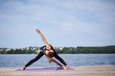 Çıplak ayaklı genç esmer kadın, siyah ve mor spor kıyafeti giyiyor, yaz aylarında tahta iskelede menekşe yoga minderinin üzerinde geriniyor. Göl kenarında yoga pozları veren formda bir kız. Sağlıklı yaşam tarzı.