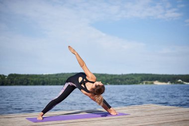 Çıplak ayaklı genç esmer kadın, siyah ve mor spor kıyafeti giyiyor, yaz aylarında tahta iskelede menekşe yoga minderinin üzerinde geriniyor. Göl kenarında yoga pozları veren formda bir kız. Sağlıklı yaşam tarzı.