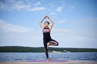 Çıplak ayaklı genç esmer kadın, siyah ve mor spor kıyafeti giyiyor, yaz aylarında tahta iskelede menekşe yoga minderinin üzerinde geriniyor. Göl kenarında yoga pozları veren formda bir kız. Sağlıklı yaşam tarzı.
