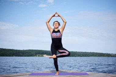 Çıplak ayaklı genç esmer kadın, siyah ve mor spor kıyafeti giyiyor, yaz aylarında tahta iskelede menekşe yoga minderinin üzerinde geriniyor. Göl kenarında yoga pozları veren formda bir kız. Sağlıklı yaşam tarzı.
