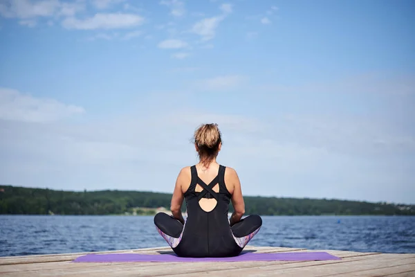 Young brunette woman, wearing black and purple fitness outfit, sitting on violet yoga mat outside on wooden pier in summer. Fit girl, doing yoga poses by lake, thinking, relaxing, meditating