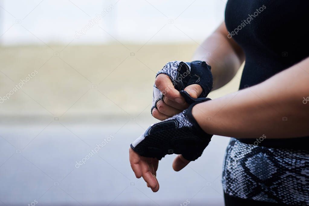 Close-up picture of hands, putting on and fastening black fitness gloves. Sportswoman, preparing for the training outside. Healthy lifestyle concept.