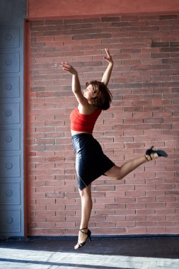 Genç esmer kadın, kırmızı bluzlu ve siyah deri etekli, geniş bir stüdyo dairede büyük bir pencereyle dans ediyor. Bachata salsa dansçısı, tuğla duvarlı bir apartman dairesinde eğitim görüyor..