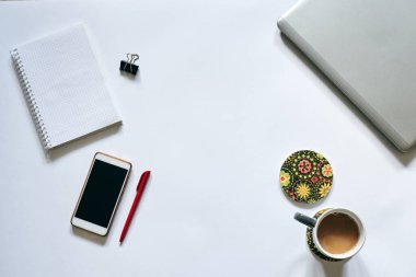 Beyaz arka planda izole edilmiş ofis aksesuarlarının düz tablosu. Gri metalik dizüstü bilgisayar, beyaz akıllı telefon, kırmızı kalem, cep telefonu defteri ve masada gri kahve bardağı. İnternette çalışıyorum. Evde kal..
