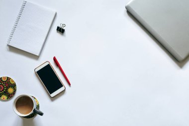Beyaz arka planda izole edilmiş ofis aksesuarlarının düz tablosu. Gri metalik dizüstü bilgisayar, beyaz akıllı telefon, kırmızı kalem, cep telefonu defteri ve masada gri kahve bardağı. İnternette çalışıyorum. Evde kal..