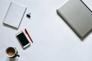 Beyaz arka planda izole edilmiş ofis aksesuarlarının düz tablosu. Gri metalik dizüstü bilgisayar, beyaz akıllı telefon, kırmızı kalem, cep telefonu defteri ve masada gri kahve bardağı. İnternette çalışıyorum. Evde kal..