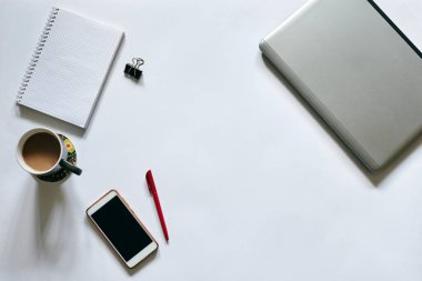 Beyaz arka planda izole edilmiş ofis aksesuarlarının düz tablosu. Gri metalik dizüstü bilgisayar, beyaz akıllı telefon, kırmızı kalem, cep telefonu defteri ve masada gri kahve bardağı. İnternette çalışıyorum. Evde kal..