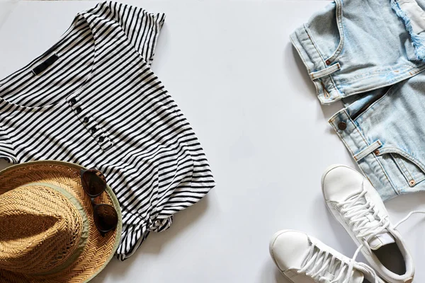 Flat lay picture of summer clothes, isolated on white background. Straw hat, sunglasses, stripy black and white t-shirt, light blue jeans shorts and white sneakers. Summer vacation background.