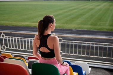 Genç esmer sporcu kadın, siyah bluz, pembe tayt ve spor ayakkabı giyiyor, stadyum tribününde renkli koltuklarda oturuyor, antrenmandan sonra dinleniyor, düşünüyor. Etkin sağlıklı yaşam tarzı konsepti.