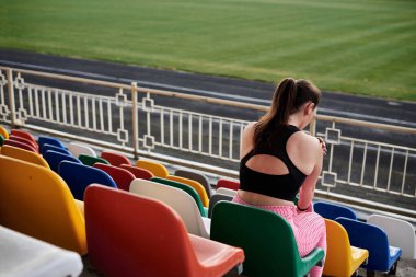 Genç esmer sporcu kadın, siyah bluz, pembe tayt ve spor ayakkabı giyiyor, stadyum tribününde renkli koltuklarda oturuyor, antrenmandan sonra dinleniyor, düşünüyor. Etkin sağlıklı yaşam tarzı konsepti.