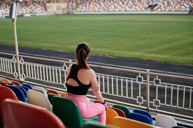 Genç esmer sporcu kadın, siyah bluz, pembe tayt ve spor ayakkabı giyiyor, stadyum tribününde renkli koltuklarda oturuyor, antrenmandan sonra dinleniyor, düşünüyor. Etkin sağlıklı yaşam tarzı konsepti.