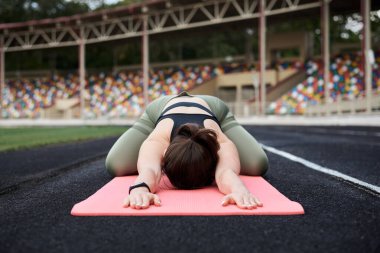 Genç esmer sporcu, siyah üst ve yeşil tayt giyiyor, yaz sabahı stadyumda yoga pozu veriyor, pembe yoga minderinin üzerinde antrenman yapıyor. Sağlıklı aktif yaşam konsepti.