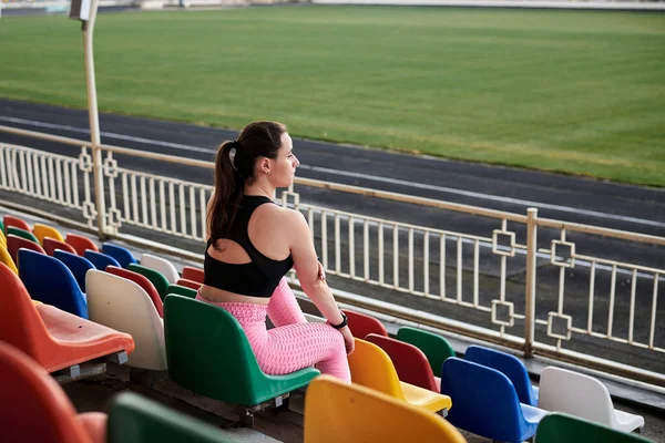 年轻的黑发女运动员 身穿黑色上衣 粉红的腿和运动鞋 坐在体育场讲台上五颜六色的座位上 经过训练和思考后放松了下来 积极健康的生活方式概念 — 图库照片
