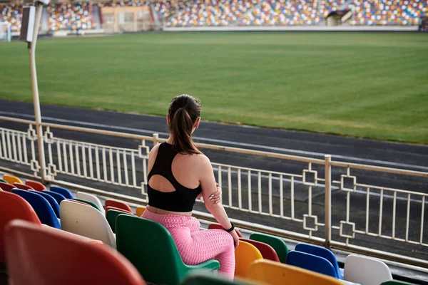年轻的黑发女运动员 身穿黑色上衣 粉红的腿和运动鞋 坐在体育场讲台上五颜六色的座位上 经过训练和思考后放松了下来 积极健康的生活方式概念 — 图库照片