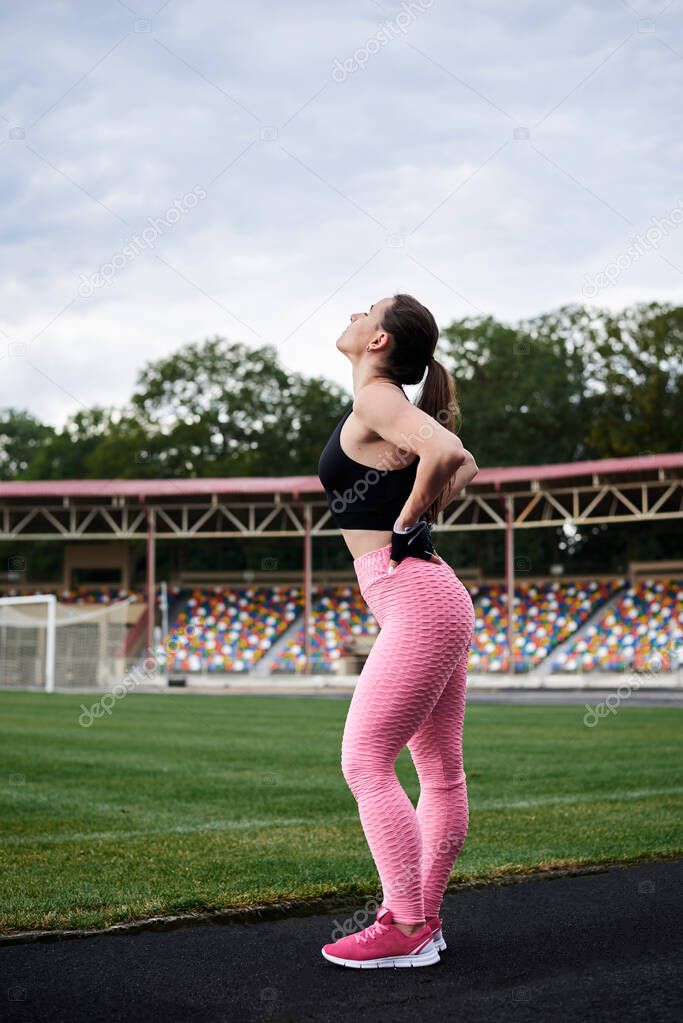 Young brunette woman, wearing pink leggings, sneakers, black top and fitness gloves, training on city stadium in summer. Sportswoman, stretching, warming up before exercise outside. Healthy lifestyle