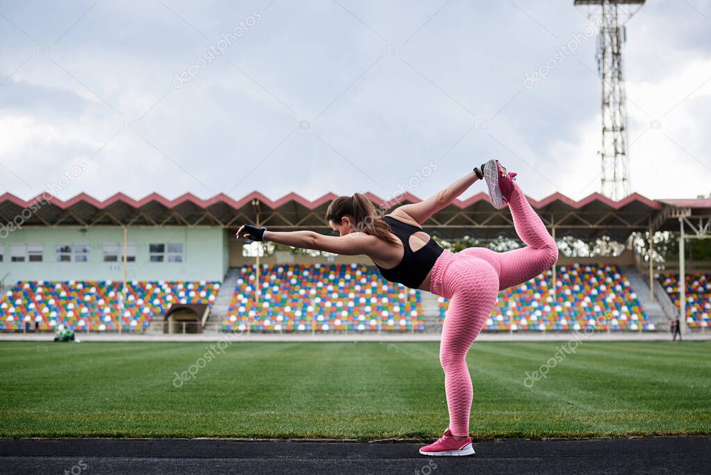 Young brunette woman, wearing pink leggings, sneakers, black top and fitness gloves, training on city stadium in morning. Sportswoman, stretching, warming up before exercise outside. Healthy lifestyle
