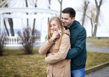 Genç esmer adam, yeşil ceket giyiyor, genç sarışın kadının arkasında duruyor, bej ceket giyiyor, ona sarılıyor, gülümsüyor, öpüşüyor. Romantik Sevgililer Günü Kutlaması