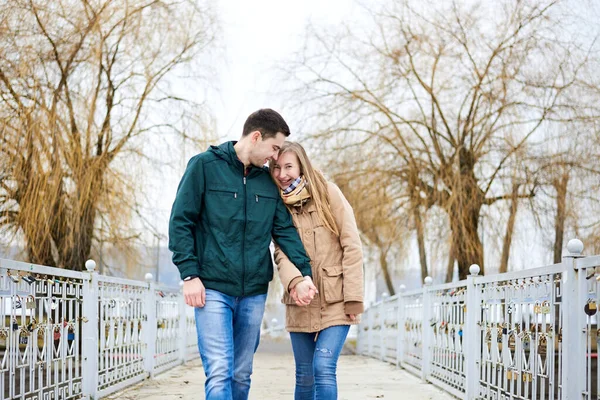年轻夫妇恋爱了 穿着米黄色和绿色休闲装 手牵手 冬天走在锁桥上 背景是黄树 情人节浪漫的庆祝活动 — 图库照片