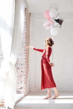 Kısa kıvırcık saçlı, kırmızı elbiseli, dans eden, pembe ve gümüş balonları tutan, büyük pencerenin yanındaki beyaz arka planda izole genç, esmer bir kadının tam boy portresi.